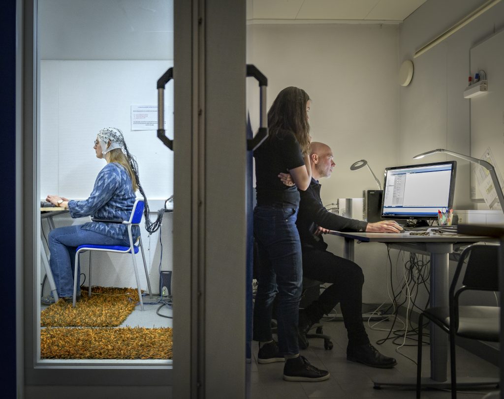 Looking at brain activity: subject on left has cap with electrodes and wires coming from it. On the other side of the wall are researchers looking at a computer. Subject and researchers are facing in opposite directions. 